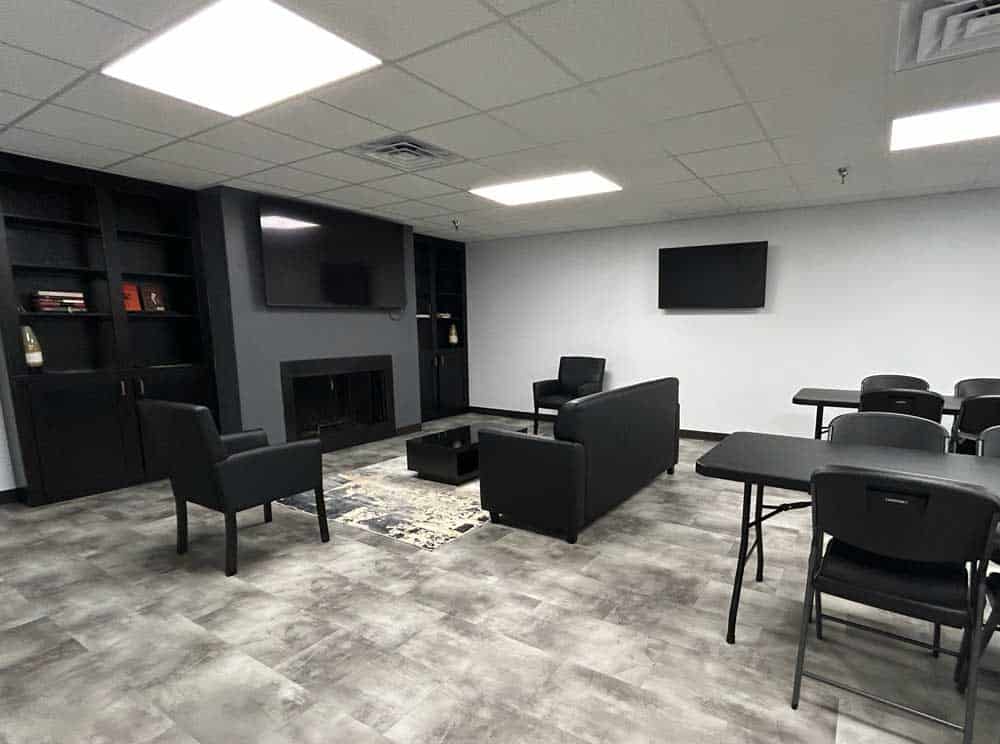 Freedom Firearms & Defense | A modern room with a seating area featuring black chairs and a sofa, a television on the wall, shelves, and several tables and chairs. The floor is tiled, and the walls are white.