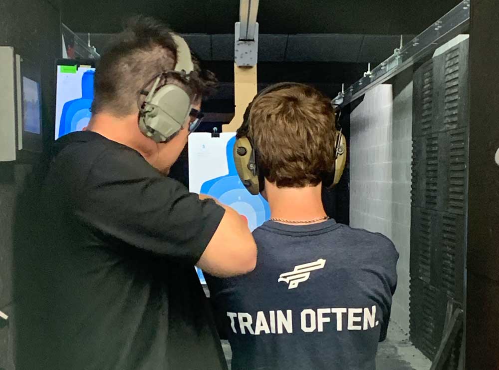 Freedom Firearms & Defense | Two people wearing ear protection are at a shooting range, aiming at a target. The person on the right is wearing a shirt that says "TRAIN OFTEN.